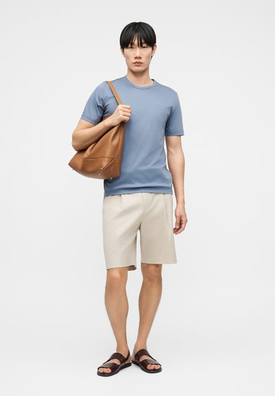 Νέος άνδρας που φοράει μπλε μπλουζάκι, μπεζ σορτς, καφέ σανδάλια, κρατώντας μια μεγάλη καφέ δερμάτινη τσάντα ώμου μπροστά σε απλό φωτεινό φόντο.