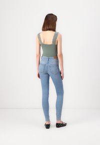 Une combinaison verte avec des bretelles en dentelle, associée à un jean skinny bleu clair. Le jean présente une taille haute et des poches arrière ; les chaussures sont noires avec des accents.