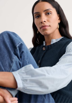 Tumši zils tamborēts vestes ar ribotiem akcentiem un pogas aizdari, valkāts kopā ar gaiši zilu rievotu blūzi ar volāniem; modelis sēž džinsa biksēs.