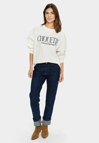 Tejfehér pulóver fekete "CHOUETTE" felirattal, sötétkék farmerrel, ami fel van rolled a száraknál, és barna velúrbakancsos bokacsizmával.