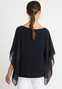 Svart blus med genomskinliga batwingärmar, en vid halsringning och ett lätt material. Utformad för en lös och flödande passform.