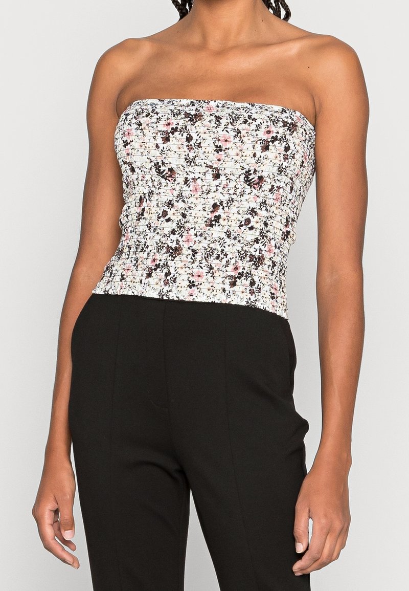 Femme portant un haut blanc sans bretelles, ajusté, avec un petit imprimé floral noir et rose, associé à un pantalon noir taille haute sur un fond uni.