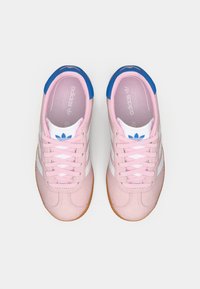 Um par de ténis Adidas rosa com riscas brancas, detalhes em azul no calcanhar, atacadores rosa claro e soles cor de goma sobre um fundo branco.