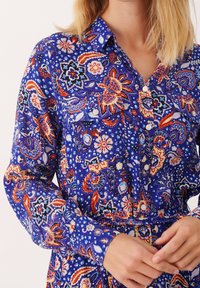 Niebieska koszula z kwiatowymi i paisleyowymi wzorami w kolorze czerwonym, białym i czarnym. Posiada zapięcie na guziki, kołnierzyk i długie rękawy.