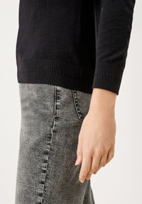Schwarzer Strickpullover mit geripptem Saum, kombiniert mit grauen Jeans aus Denim mit verblasster Textur und kontrastierenden Nähten. Eine Hand ruht auf den Jeans.