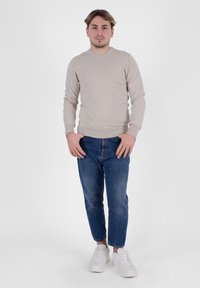 Camicia a maniche lunghe di un colore beige chiaro con una texture a coste, abbinata a jeans blu slim-fit e sneaker bianche, in piedi contro uno sfondo semplice.