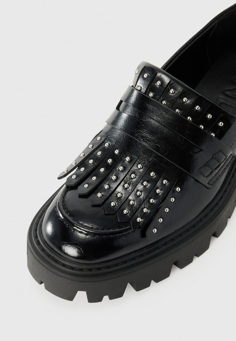 Mocassino in pelle nera con dettagli di frange chiodate, suola nera chunky e finitura lucida, che mette in risalto la texture e il design.