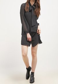 Abito nero a pois con maniche lunghe e un fiocco al collo, sovrapposto a una gonna più corta. La modella indossa stivaletti neri alla caviglia.