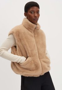 Beige-farbenes Kunstfell-Weste ohne Ärmel mit hohem Kragen, Reißverschluss und zwei Seitentaschen, getragen über einem hellbeigen Langarmtop.