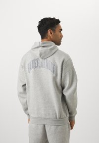 Sudadera gris con una textura suave, que cuenta con capucha y un destacado logo de "UNDER ARMOUR" en azul en la parte posterior, puños y dobladillo acanalados.