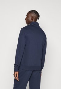 Μπλε ναυτικό φούτερ με κοντό φερμουάρ, ψηλό γιακά και ριμπ μανσέτες. Μαλακό υλικό με λεία υφή, σχεδιασμένο για εφαρμοστή εμφάνιση.