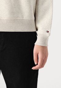 Sweat-shirt beige clair avec des poignets et un ourlet côtelés. Petite patch logo sur le poignet. Porte avec un pantalon en denim noir. Texture douce.
