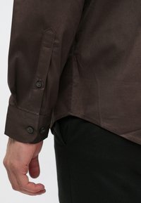 Mørkebrun langærmet skjorte med knapper ved ærmegabene. Fremstillet af glat stof, med en skræddersyet pasform og en buet nederkant.