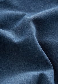 Tummansininen farkkukangas, jossa on sileä, hieman teksturoitu pinta. Materiaali on ryppyinen, mikä korostaa sen joustavaa kaartoa ja värin syvyyttä.