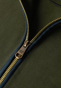 Kurtka polarowa zapinana na zamek błyskawiczny w ciemnozielonym kolorze z granatowymi akcentami. Posiada mosiężny zamek z grawerowanym detalem. Gładka, miękka struktura na całej powierzchni.