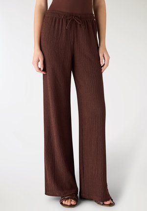 Donna che indossa pantaloni marroni larghi e strutturati con vita a coulisse e sandali marroni, in piedi contro uno sfondo bianco semplice.