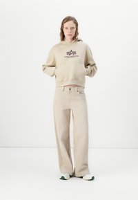 Donna che indossa un felpa beige di Alpha Industries, pantaloni beige a gamba larga e sneakers bianche, in piedi su uno sfondo bianco uniforme.