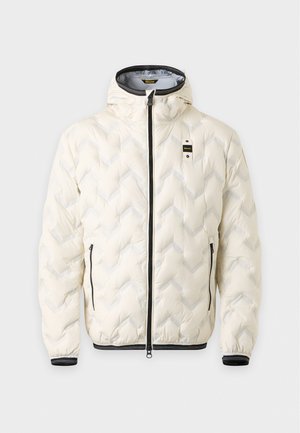 Un homme porte une veste blanche matelassée à capuche, un pantalon noir et des baskets blanches sur un fond clair uni.