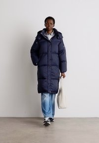 Giacca puffer blu navy con cappuccio, bottoni a pressione e zip laterali; abbinata a jeans azzurri e sneakers nere, mentre porta una borsa tote bianca.