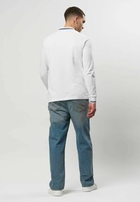 Homme portant un polo à manches longues blanc avec un col rayé, un jean bleu et des baskets blanches, debout, de dos, sur un fond uni.
