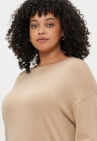 Μπεζ πλεκτό πουλόβερ με στρογγυλή λαιμόκοψη και άνετη εφαρμογή. Διαθέτει ριγωτή υφή και χαμηλούς ώμους. Επισημαίνεται από διακριτές ραφές.