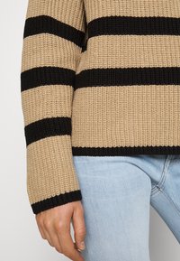 Beige gebreide trui met zwarte horizontale strepen; heeft een textuurrijke stof en geribde manchetten, gecombineerd met lichtblauwe jeans.