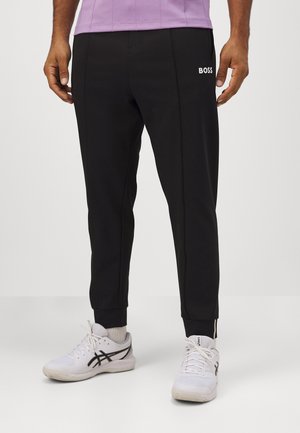 Black jogger pants with a slim fit, featuring a white "BOSS" logo and zipper details at the ankles. Made from smooth, stretchy fabric.