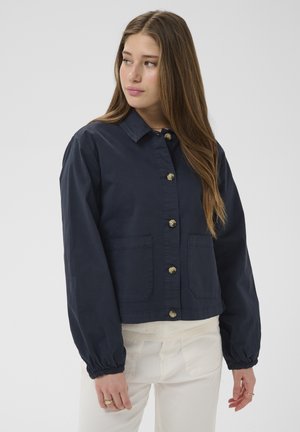 Noor pruuni pikkade juustega naine, kes kannab tumesinist nööpidega jopet ja valgeid pükse, vaatab vasakule, taust on lihtne.