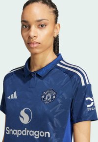 Maillot bleu de Manchester United avec un col. Comprend des bandes blanches Adidas sur les épaules, un logo et le nom du sponsor sur le devant.