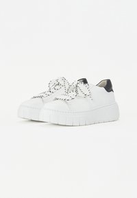 Sneaker bianco con suola spessa, lacci intrecciati bianchi e neri, tomaia in pelle e un accento nero sul tallone. Design della punta leggermente arrotondato.