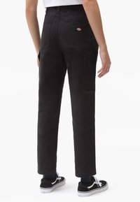 Persona que lleva pantalones negros de pierna recta con bolsillos de parche y un logo en el bolsillo trasero, combinados con zapatillas negras y blancas.