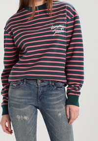 Sudadera a rayas verdes y rosas con cuello redondo y un texto de logo "tommy jeans" en el pecho, combinada con jeans azules desgastados.