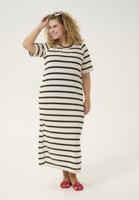 Gestreiftes Maxikleid in Creme und Schwarz, mit kurzen Ärmeln, rundem Halsausschnitt und einem seitlichen Schlitz; getragen mit roten Sandalen und accessorized mit einer Brille.