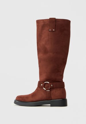 Brown suede knee-high boot with a flat sole, a buckle strap around the ankle, and a rounded toe. Smooth texture and simple design.