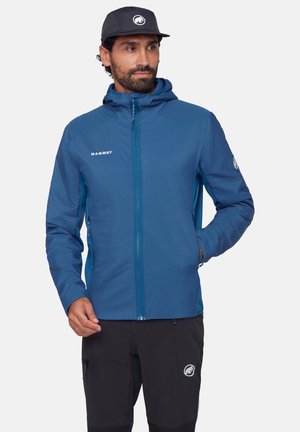 Uomo con giacca blu Mammut con cappuccio, pantaloni neri e cappellino, che guarda di lato con una mano in tasca e l'altra che sistema la giacca.
