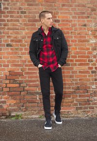 Schwarze Jeansjacke über einem rot-schwarzen karierter Hemd, kombiniert mit schwarzen Hosen und schwarzen High-Top Sneakern, vor einer Ziegelmauer.
