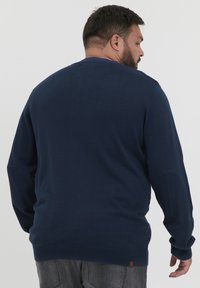 Jersey azul marino con un cuello y puños acanalados, fabricado en tejido de punto. Presenta un pequeño acento de cuero rectangular en el dobladillo.
