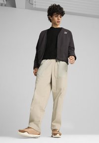 Un outfit da uomo composto da una giacca grigia scura con zip, un maglione nero a texture, pantaloni beige a gamba larga con accenti chiari e sneakers marroni.