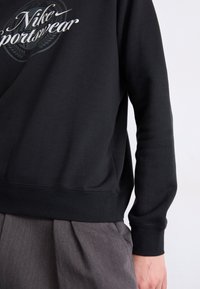 Črna kapuca s belim logotipom "Nike Sportswear", z ribanim robom in manšetami. Združena z sivimi plisejskimi hlačami.