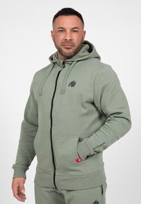 Hoodie in gedempt groen, met een volledige voorkantrits, kangoeroezakken en een zwart gorilla-logo op de borst. Van een katoenmix.