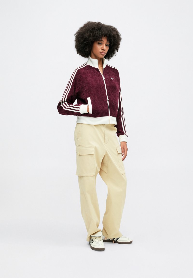 Bordeaux velours zip-up jas met witte accenten, gecombineerd met beige cargobroek. De jas heeft ribbelboorden en drie witte strepen op de mouwen.