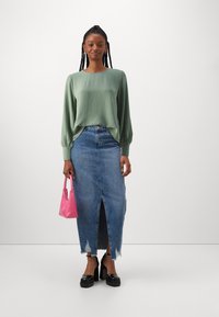 Top de manga larga texturizada verde combinado con una falda de mezclilla con una abertura frontal y dobladillo desgastado. Accesorios con un pequeño bolso rosa.