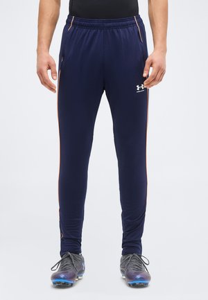 Άνδρας που φοράει αθλητικό παντελόνι σκούρου μπλε με πορτοκαλί ρίγες στο πλάι και αθλητικά παπούτσια, στέκεται με το πρόσωπο προς τα εμπρός σε απλό φόντο.