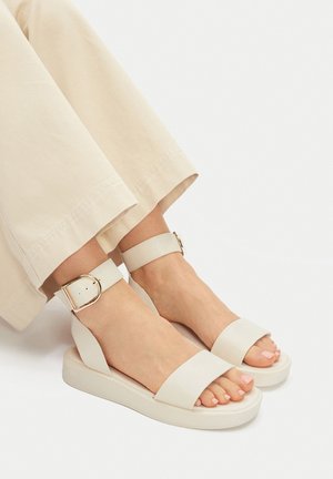 Białe skórzane sandały na platformie z szerokimi paskami i klamrą przy kostce. Gumowa podeszwa. Czysty design, gładka faktura i minimalistyczny styl.