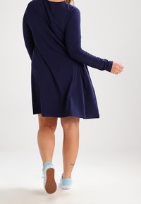 Granatowa sukienka z długimi rękawami, z drapowanym tyłem i rozkloszowanym dołem. Para jasnoniebieskich sneakersów z teksturowaną podeszwą.