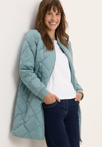 Giacca trapuntata azzurra chiara con chiusura a zip, polsini a costine e tasche laterali, indossata sopra un top bianco e jeans blu navy.