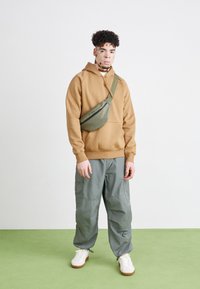 Brown hooded sweatshirt, olive green fanny pack with a zipper, gray cargo pants, and white sneakers with brown soles. Background is light gray.