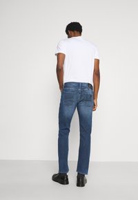 Man i vit t-shirt, blå jeans och svarta skor som står vänd med ryggen mot en enkel vit bakgrund.