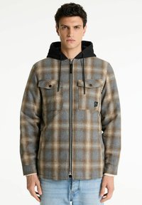 Karierte Flanelljacke in Grau-, Braun- und Cremetönen, ausgestattet mit einer schwarzen Kapuze, zwei Brusttaschen und einem Reißverschluss vorne.