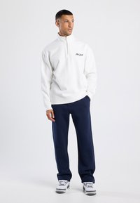 Felpa bianca a zip con collo a quarti con "New York" ricamato sul petto, abbinata a pantaloni blu scuro e sneakers bianche. Tessuto dal tocco morbido.